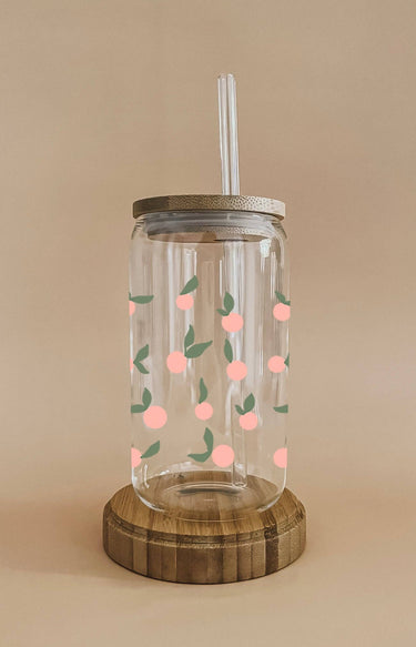 A clear glass tumbler with a bamboo lid and straw features a pattern of light orange peach decals with green leaves and rests on a round wooden coaster.